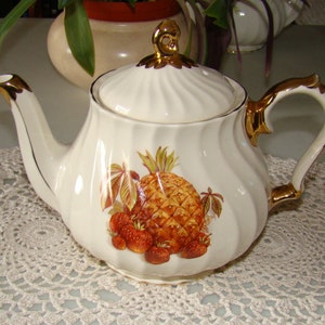 May include: White teapot with a gold trim and a pineapple and strawberry design. The teapot has a lid with a gold knob.