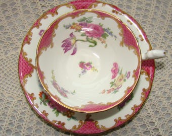 May include: A pink and gold teacup and saucer set with a floral pattern. The cup and saucer have a scalloped edge and a gold trim.