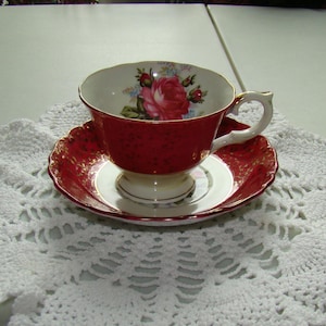 May include: A red teacup and saucer set with a floral design inside the cup. The cup and saucer have a gold trim. The cup has a white handle. The saucer is round with a scalloped edge. The set is on a white crocheted doily.