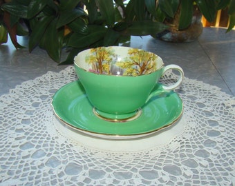 May include: A vintage green teacup and saucer set. The cup and saucer are a vibrant green with gold trim. The inside of the cup features a scenic landscape design. The set is placed on a white lace doily.