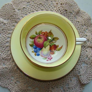 May include: A yellow teacup and saucer set with a gold rim. The teacup features a painted design of fruit, including a pear, apple, and grapes.