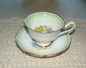 May include: A white teacup and saucer with a green and gold rim. The cup and saucer are decorated with a floral pattern of yellow and blue flowers.