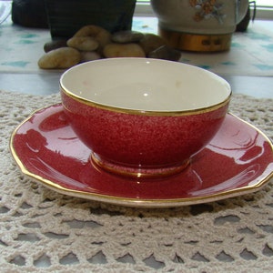 May include: A red teacup and saucer with a gold rim. The saucer has a scalloped edge. The teacup and saucer are on a white crocheted doily.
