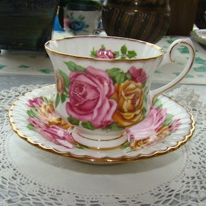 May include: A white teacup and saucer with a gold rim and floral design. The design features pink and yellow roses with green leaves.