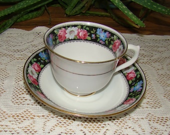 May include: A white teacup and saucer set with a black floral border featuring pink, white, and blue flowers. The teacup and saucer have gold trim. The teacup is sitting on the saucer.
