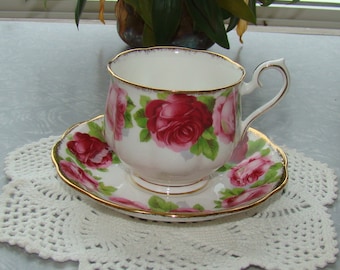 May include: A white teacup and saucer with a gold rim and a floral pattern of pink roses. The teacup and saucer are on a white crocheted doily.