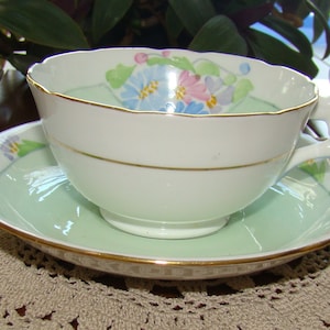 May include: White teacup and saucer set with a light green interior and gold trim. The cup features a floral design with blue and pink flowers. The saucer has a matching floral pattern. A classic tea set.