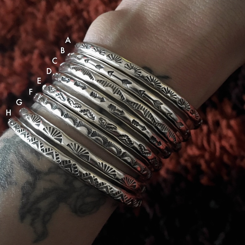 K&ouml;nnte beinhalten: Ein Set aus acht silbernen Armb&auml;ndern mit verschiedenen ge&auml;tzten Designs, darunter Pfeile, Sonnenstrahlen und Wellenlinien. Die Armb&auml;nder sind auf dem Handgelenk einer Person gestapelt.