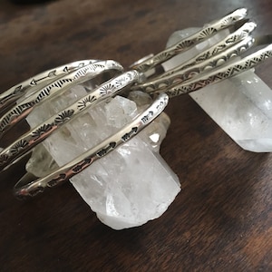 K&ouml;nnte beinhalten: Ein Set aus f&uuml;nf silbernen Armb&auml;ndern mit verschiedenen ge&auml;tzten Designs. Die Armb&auml;nder sind &uuml;bereinander gestapelt und liegen auf einem klaren Quarzkristall.