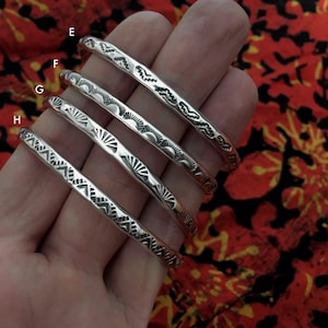 K&ouml;nnte beinhalten: Vier silberne Armb&auml;nder mit verschiedenen gravierten Designs. Die Armb&auml;nder sind &uuml;bereinander gestapelt, wobei das oberste Armband ein wellenf&ouml;rmiges Design hat, das zweite Armband ein Sonnenstrahl-Design, das dritte Armband ein Sonnenstrahl-Design und das unterste Armband ein geometrisches Design.