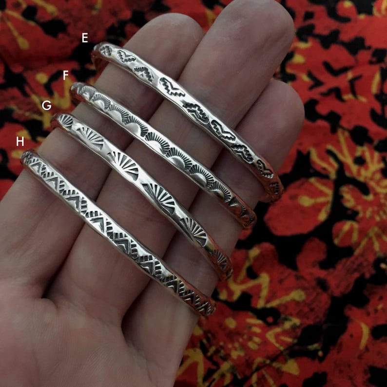 K&ouml;nnte beinhalten: Vier silberne Armb&auml;nder mit verschiedenen gravierten Designs. Die Armb&auml;nder sind &uuml;bereinander gestapelt, wobei das oberste Armband ein wellenf&ouml;rmiges Design hat, das zweite Armband ein Sonnenstrahl-Design, das dritte Armband ein Sonnenstrahl-Design und das unterste Armband ein geometrisches Design.