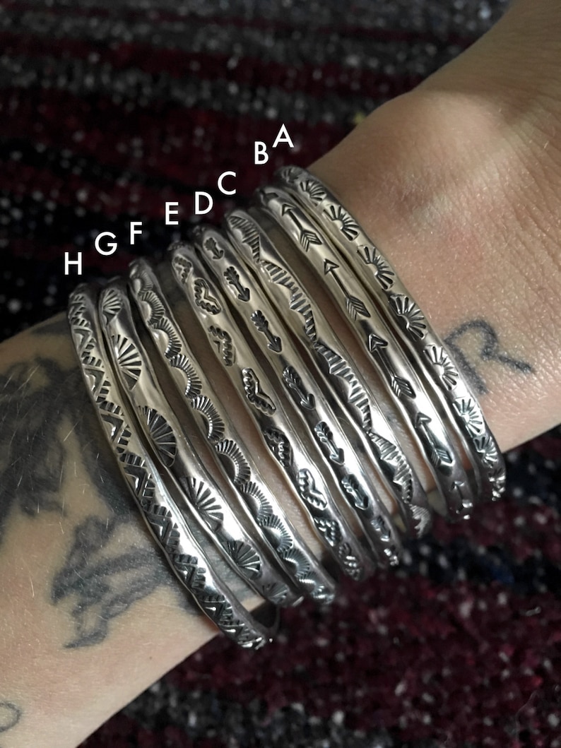 K&ouml;nnte beinhalten: Acht silberne Armb&auml;nder mit verschiedenen ge&auml;tzten Designs. Die Armb&auml;nder sind auf dem Handgelenk einer Person gestapelt.
