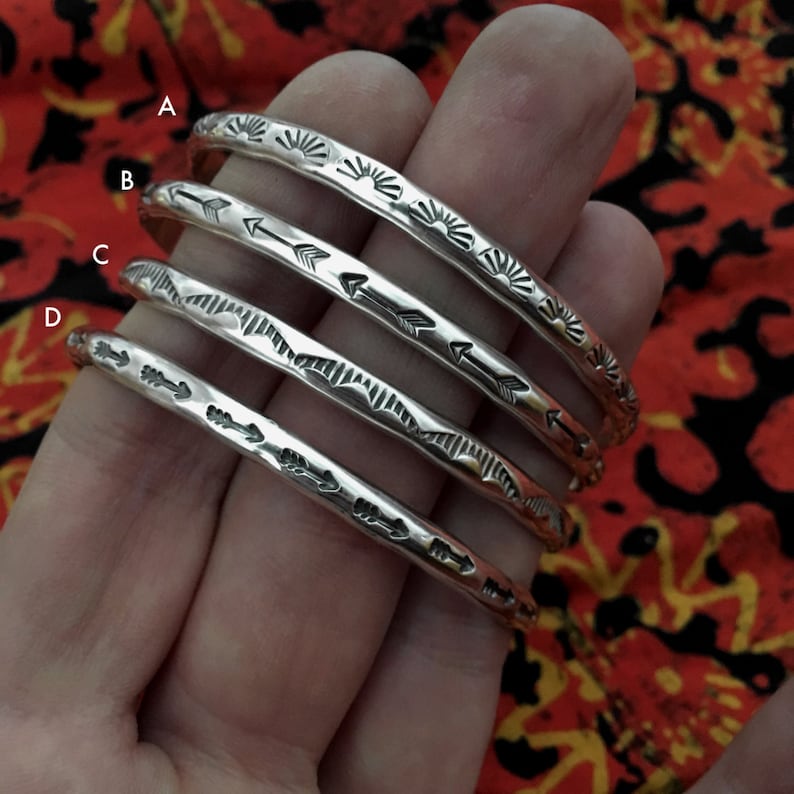 K&ouml;nnte beinhalten: Vier silberne Armb&auml;nder mit verschiedenen Designs. Das obere Armband hat ein Sonnenstrahlenmuster, das zweite hat Pfeile, das dritte hat ein geometrisches Muster und das untere Armband hat Pfeile.