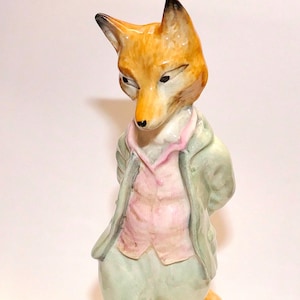 May include: A ceramic figurine of an anthropomorphic fox standing on a green base. The fox has orange fur and is wearing a light green jacket, a pink vest, and light green breeches. The figure is painted with a glossy finish and fine details.