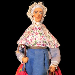 May include: A vintage doll dressed in traditional attire, holding a red umbrella and a small copper bucket. The doll wears a floral patterned shawl, blue pants, and a red skirt with a yellow and green floral design. The doll's face is painted with a serene expression.