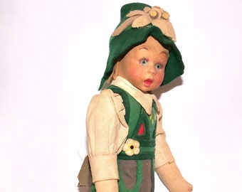 May include: A vintage cloth doll wearing a green and white hat with a flower design, a white shirt, and brown shorts with green trim. The doll has a painted face with blue eyes and a pink complexion.