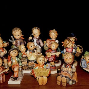 May include: A collection of Hummel figurines depicting children in various activities. The figurines are painted in muted colors and feature children playing musical instruments, carrying baskets, and dressed in traditional clothing. An ashtray is also present.
