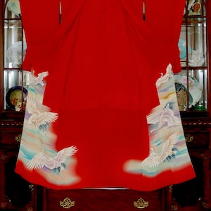 May include: A red kimono with white cranes and clouds embroidered on the sleeves and hem. The kimono is hanging on a wooden stand.