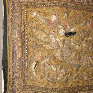 May include: An ornate textile with a detailed, gold-toned design featuring figures in traditional attire. The artwork is framed by a border of purple and gold, with a dark green edge. The textile is likely a decorative piece or wall hanging.