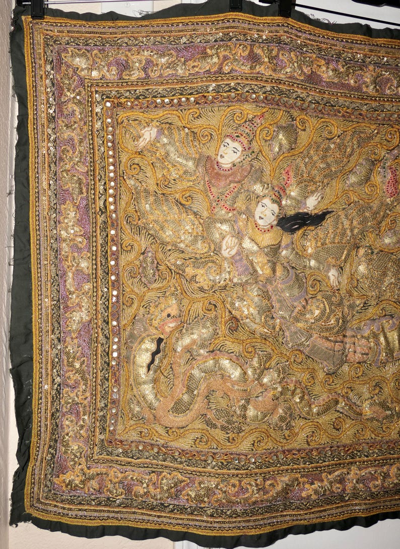 May include: An ornate textile with a detailed, gold-toned design featuring figures in traditional attire. The artwork is framed by a border of purple and gold, with a dark green edge. The textile is likely a decorative piece or wall hanging.