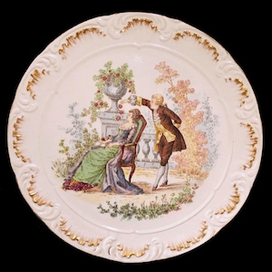 May include: A decorative porcelain plate with a scalloped edge and gold trim. The plate features a colorful scene of a man offering fruit to a seated woman in a garden setting. The plate has a cream-colored background.