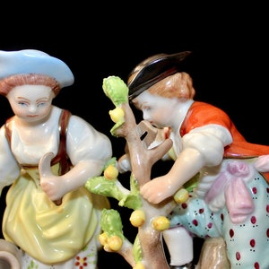Puede incluir: Un conjunto de figuras de porcelana que representa a dos personas, una mujer con un sombrero azul y un hombre con un sombrero marr&oacute;n, cuidando un peque&ntilde;o &aacute;rbol con hojas verdes y frutos amarillos. La mujer sostiene una jarra en su mano izquierda y una hoz en su mano derecha. El hombre est&aacute; alcanzando una fruta en el &aacute;rbol.