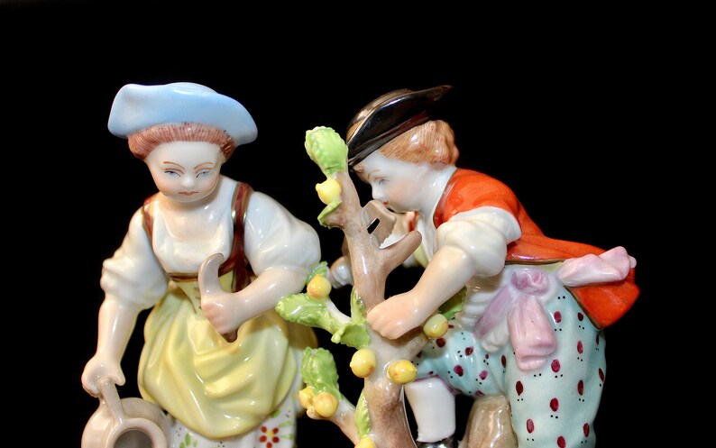 Puede incluir: Un conjunto de figuras de porcelana que representa a dos personas, una mujer con un sombrero azul y un hombre con un sombrero marr&oacute;n, cuidando un peque&ntilde;o &aacute;rbol con hojas verdes y frutos amarillos. La mujer sostiene una jarra en su mano izquierda y una hoz en su mano derecha. El hombre est&aacute; alcanzando una fruta en el &aacute;rbol.
