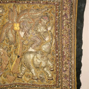 May include: An ornate textile featuring a detailed embroidered scene with gold, purple, and black threads. The central image depicts a figure riding an elephant, surrounded by intricate patterns and embellishments. The border is adorned with sequins.
