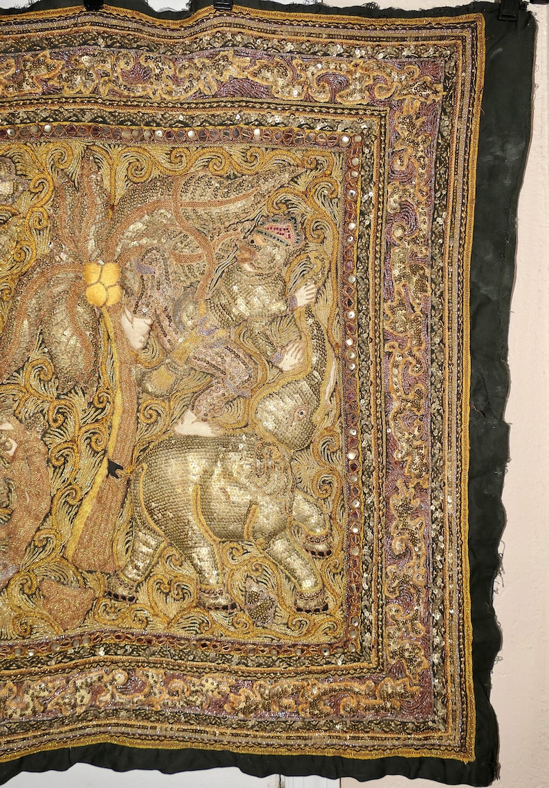 May include: An ornate textile featuring a detailed embroidered scene with gold, purple, and black threads. The central image depicts a figure riding an elephant, surrounded by intricate patterns and embellishments. The border is adorned with sequins.