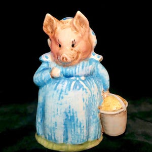 May include: A ceramic pig figurine wearing a blue and white striped dress with a matching headscarf. The pig has a pink complexion, dark eyelashes, and is holding a small, light-colored bucket in one hand. The base of the figurine is green.