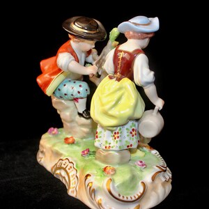 Puede incluir: Una figurita de porcelana de un ni&ntilde;o y una ni&ntilde;a con ropa tradicional. El ni&ntilde;o lleva una chaqueta roja, pantalones azules y un sombrero negro. La ni&ntilde;a lleva un corpi&ntilde;o marr&oacute;n, una falda amarilla y un sombrero azul. Est&aacute;n de pie sobre una base verde con flores.