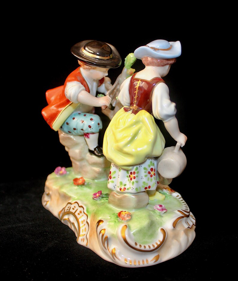 Puede incluir: Una figurita de porcelana de un ni&ntilde;o y una ni&ntilde;a con ropa tradicional. El ni&ntilde;o lleva una chaqueta roja, pantalones azules y un sombrero negro. La ni&ntilde;a lleva un corpi&ntilde;o marr&oacute;n, una falda amarilla y un sombrero azul. Est&aacute;n de pie sobre una base verde con flores.