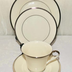 May include: Cream-colored dinnerware set with a silver trim. The set includes a dinner plate, salad plate, saucer, and teacup. The plates and cup are arranged on a white surface, showcasing the elegant design.