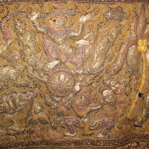 May include: Close-up of an ornate textile with intricate embroidery. The design features figures in traditional attire, with gold, brown, and pink hues. The fabric is embellished with sequins and detailed patterns.