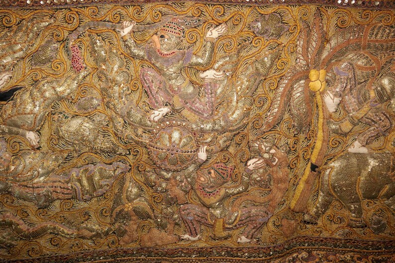 May include: Close-up of an ornate textile with intricate embroidery. The design features figures in traditional attire, with gold, brown, and pink hues. The fabric is embellished with sequins and detailed patterns.