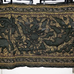 May include: Dark green quilted textile with a central design featuring stylized figures and animals in a gold and brown pattern. The border is also gold and brown. The textile is hanging from black hangers.