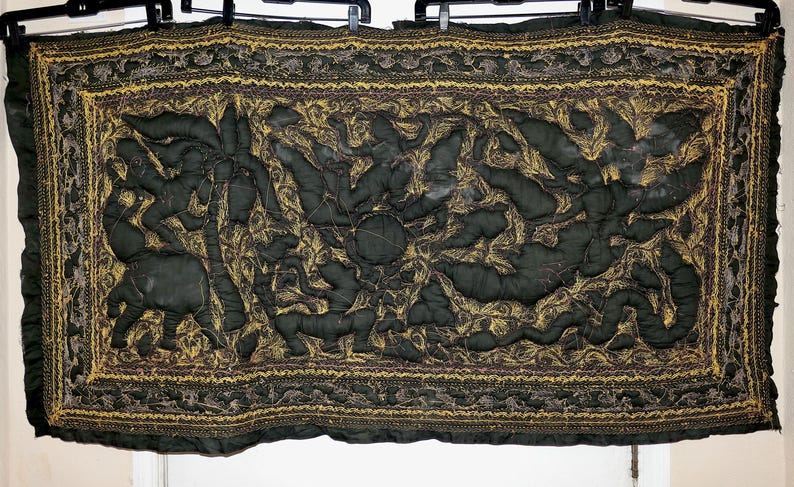 May include: Dark green quilted textile with a central design featuring stylized figures and animals in a gold and brown pattern. The border is also gold and brown. The textile is hanging from black hangers.