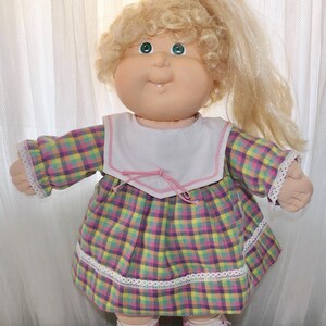 May include: A doll with blonde curly hair tied in a high ponytail, green eyes, and a single visible tooth. The doll wears a long-sleeved dress with a purple, yellow, and green plaid pattern, a white sailor-style collar, and pink plastic shoes.