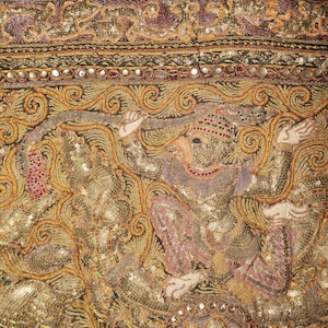 May include: Close-up of a detailed textile with ornate embroidery. The design features a figure in a seated position, with intricate gold, pink, and purple threadwork. The fabric is embellished with sequins and small mirrors, creating a shimmering effect.