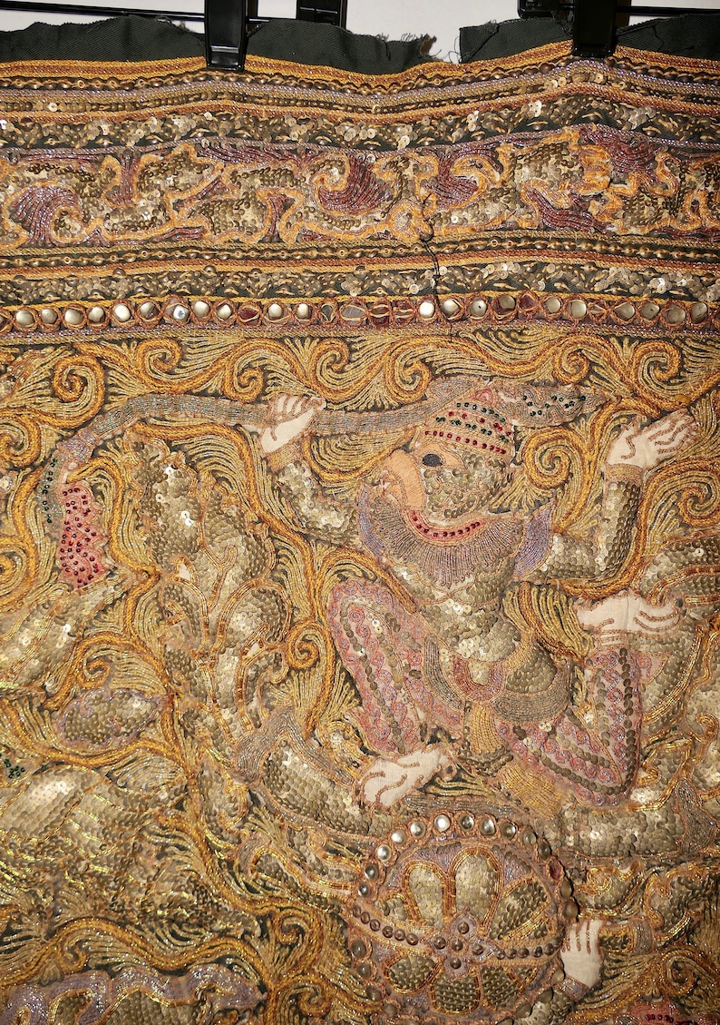 May include: Close-up of a detailed textile with ornate embroidery. The design features a figure in a seated position, with intricate gold, pink, and purple threadwork. The fabric is embellished with sequins and small mirrors, creating a shimmering effect.
