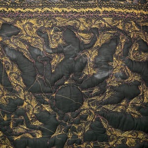 May include: Dark green quilted fabric with intricate gold and purple thread embroidery. The design features organic shapes and patterns, with a textured, handmade appearance. The fabric is held by a black hanger.