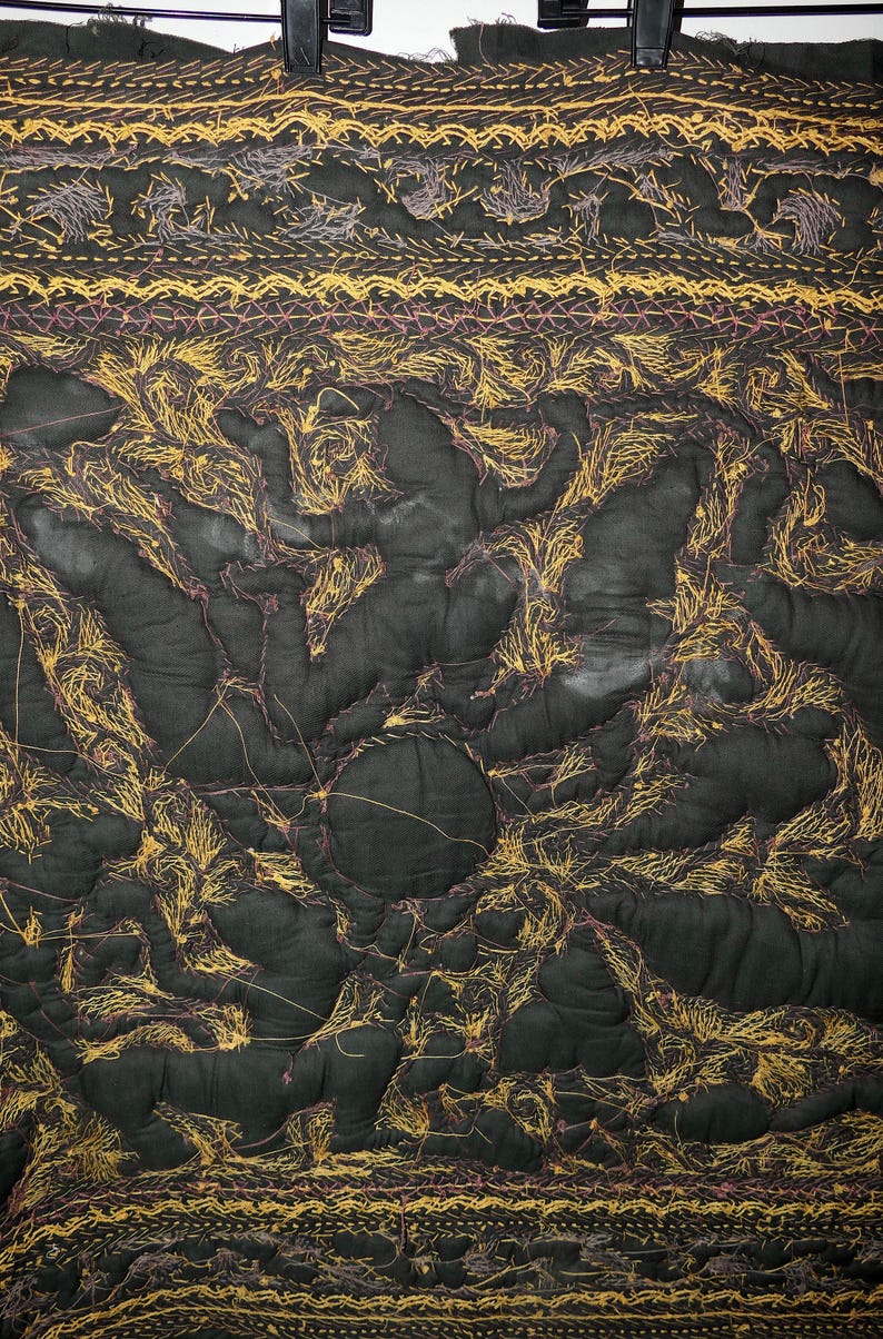 May include: Dark green quilted fabric with intricate gold and purple thread embroidery. The design features organic shapes and patterns, with a textured, handmade appearance. The fabric is held by a black hanger.