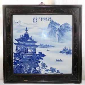 May include: A framed blue and white porcelain tile featuring a traditional Chinese landscape scene. The artwork depicts a pagoda, mountains, water, and boats. The dark wood frame has ornate carvings.