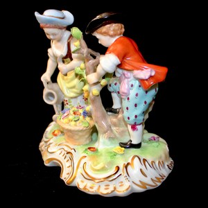 Puede incluir: Una figurita de porcelana de dos ni&ntilde;os recogiendo fruta de un &aacute;rbol. La ni&ntilde;a lleva un sombrero azul y un vestido amarillo, mientras que el ni&ntilde;o lleva una chaqueta roja y pantalones azules. La figurita est&aacute; colocada sobre una base verde y dorada.
