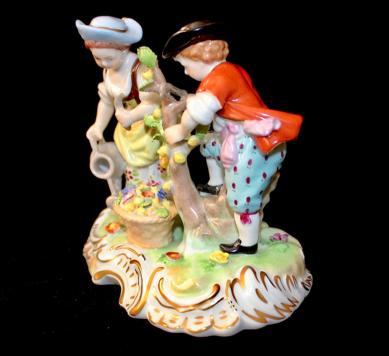 Puede incluir: Una figurita de porcelana de dos ni&ntilde;os recogiendo fruta de un &aacute;rbol. La ni&ntilde;a lleva un sombrero azul y un vestido amarillo, mientras que el ni&ntilde;o lleva una chaqueta roja y pantalones azules. La figurita est&aacute; colocada sobre una base verde y dorada.