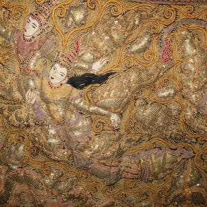 May include: Close-up of an ornate textile featuring figures in traditional attire. The figures are depicted with painted faces and elaborate headdresses, set against a backdrop of gold and brown embroidery. The textile is embellished with sequins.