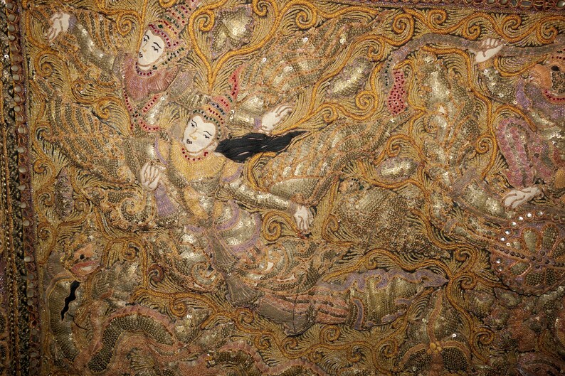May include: Close-up of an ornate textile featuring figures in traditional attire. The figures are depicted with painted faces and elaborate headdresses, set against a backdrop of gold and brown embroidery. The textile is embellished with sequins.