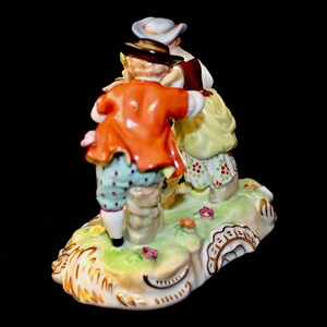 Puede incluir: Una figurita de porcelana de tres personas con ropa del siglo XVIII. La figurita est&aacute; pintada en colores brillantes, incluyendo rojo, amarillo, azul y verde. Las personas est&aacute;n de pie sobre una base verde con detalles dorados.
