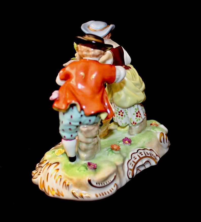 Puede incluir: Una figurita de porcelana de tres personas con ropa del siglo XVIII. La figurita est&aacute; pintada en colores brillantes, incluyendo rojo, amarillo, azul y verde. Las personas est&aacute;n de pie sobre una base verde con detalles dorados.