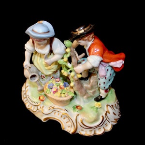 Puede incluir: Una figurita de porcelana de dos ni&ntilde;os, uno con un sombrero azul y el otro con un sombrero negro, cuidando un arbusto floreciente. La figurita est&aacute; colocada sobre una base verde y blanca con detalles dorados.